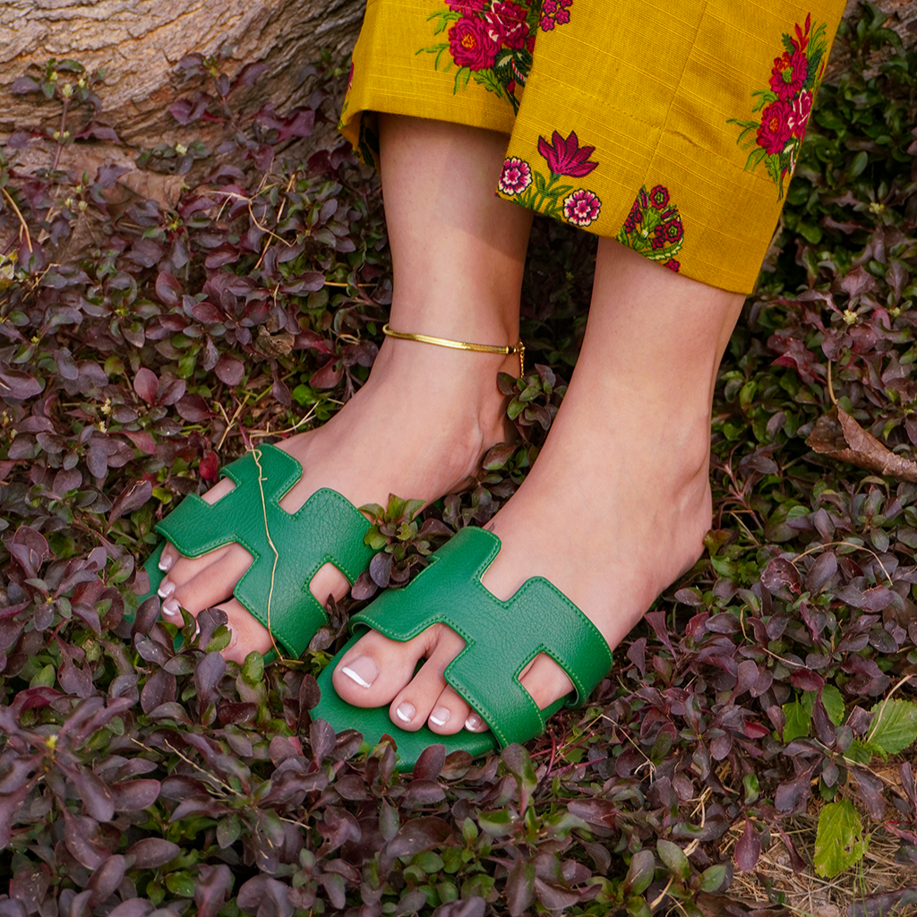 A person wearing green H style flat sandals with a cushioned sole standing on a bed of purple foliage.