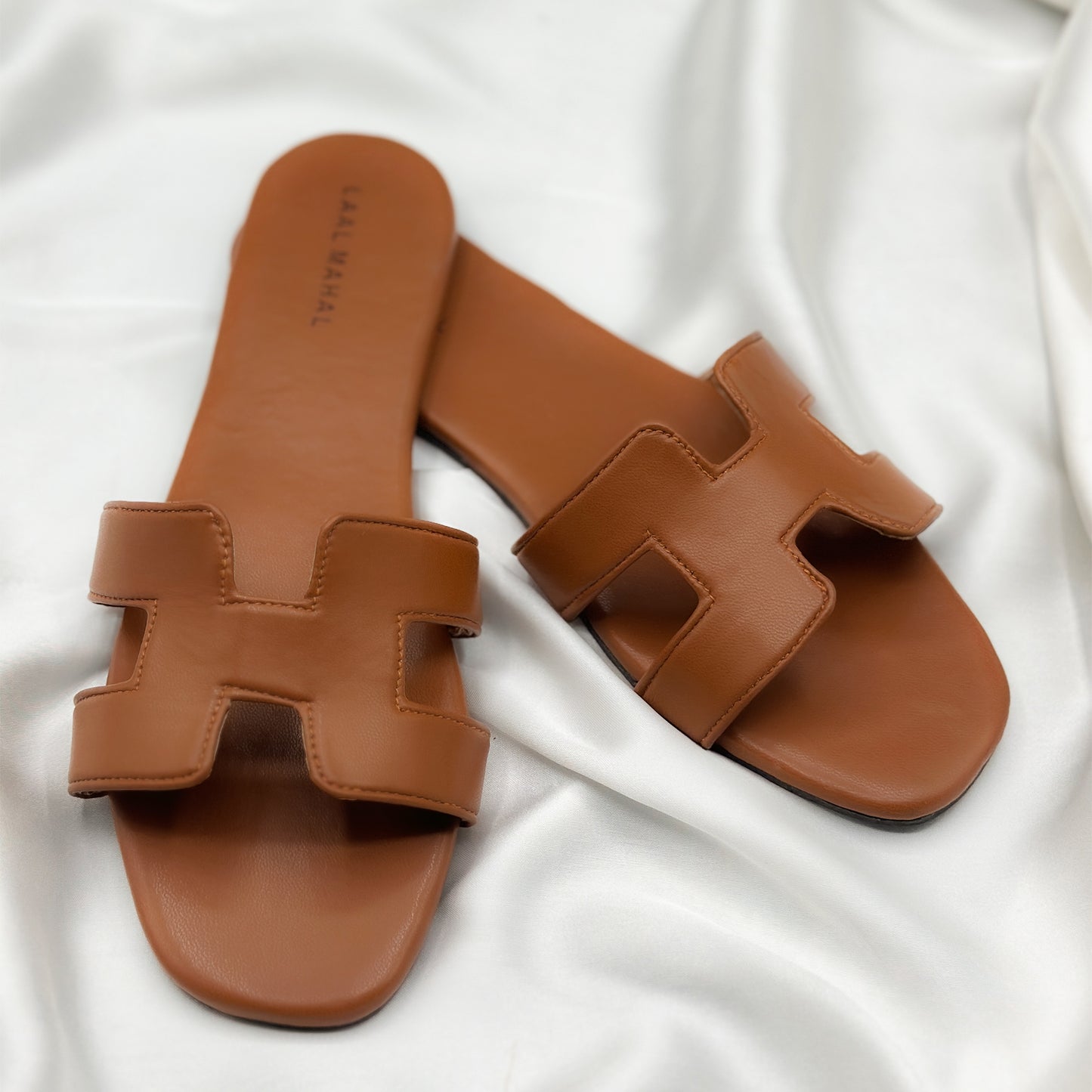 A pair of brown H-style flat sandals with a non-slip sole, placed on a white silky surface.