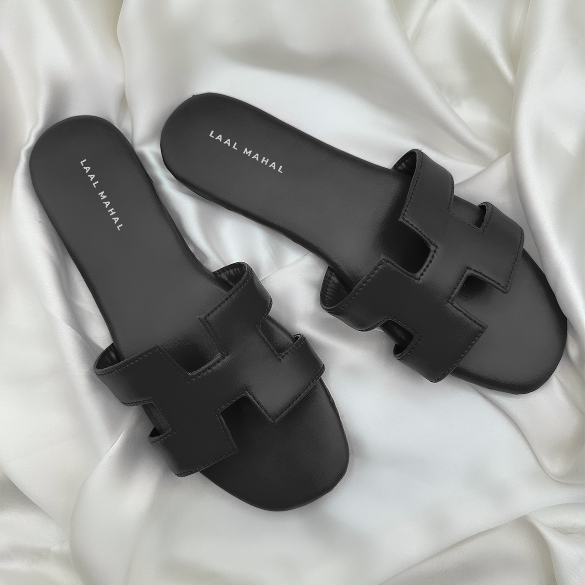 A pair of black H-style flat sandals featuring cut-out straps and a minimalist silhouette, photographed on a soft white satin background for a sleek contrast.