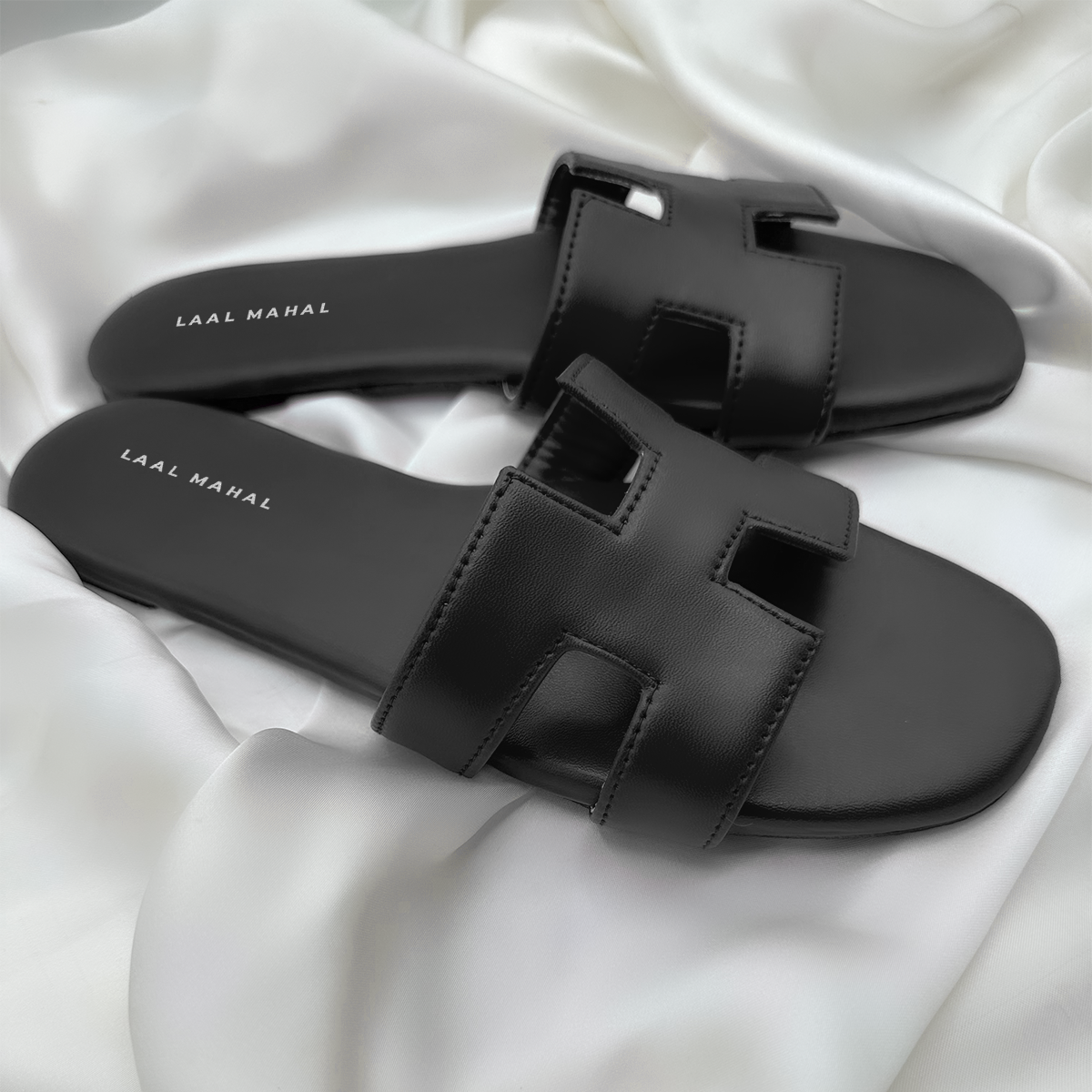 A pair of black H-style flat sandals featuring cut-out straps and a minimalist silhouette, photographed on a soft white satin background for a sleek contrast.