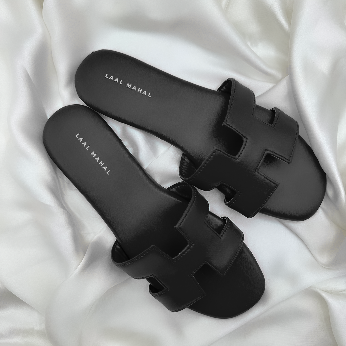 A pair of black H-style flat sandals featuring cut-out straps and a minimalist silhouette, photographed on a soft white satin background for a sleek contrast.