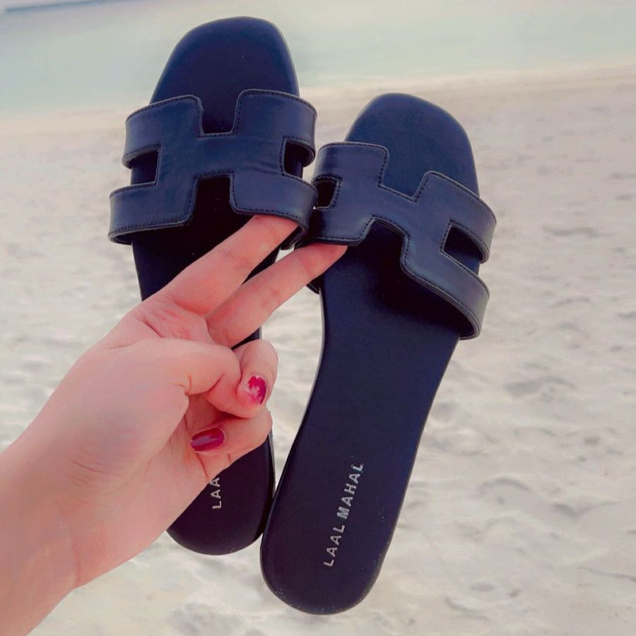 A hand holding a pair of black H-style slide sandals with cut-out straps on a sandy beach background.