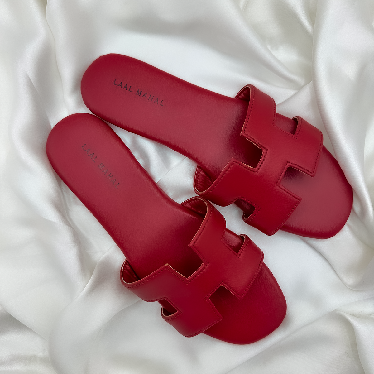 A pair of red H-style slide sandals with cut-out straps, displayed on soft white satin fabric.