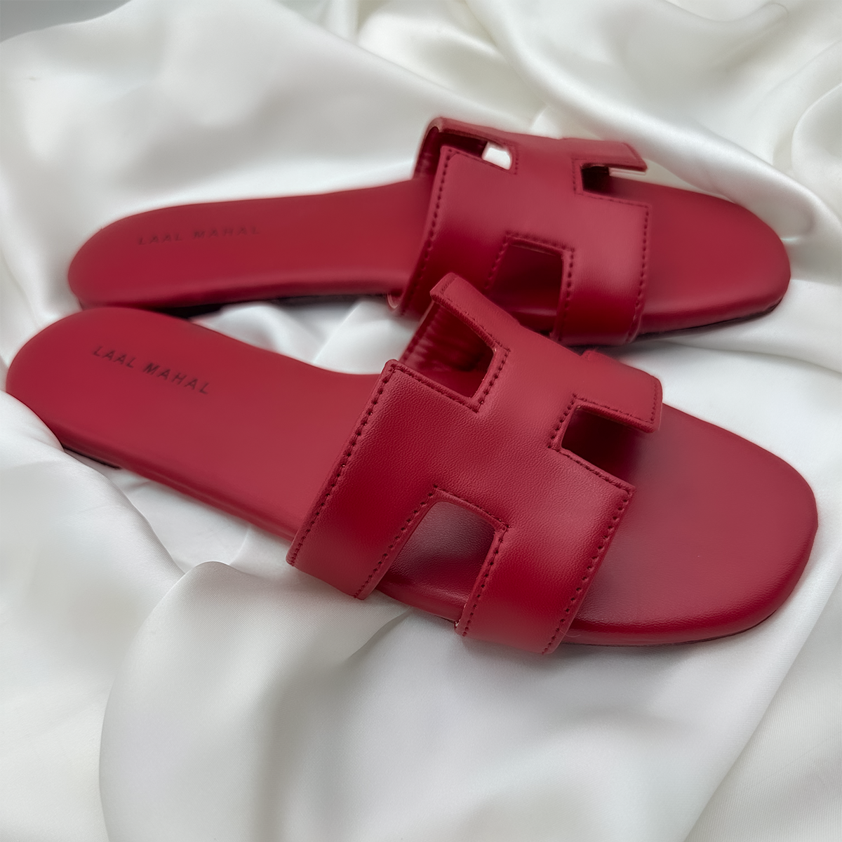 A pair of red H-style flat sandals with cut-out straps displayed on soft white satin fabric, showcasing minimalist design and smooth leather texture.