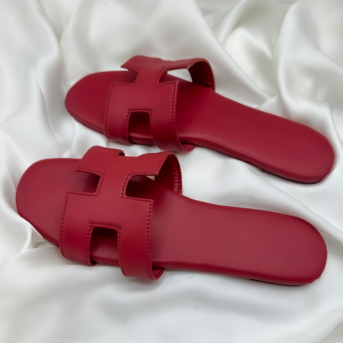 A pair of red H-style flat sandals with cut-out straps, displayed on smooth white satin fabric for a minimal and elegant look.